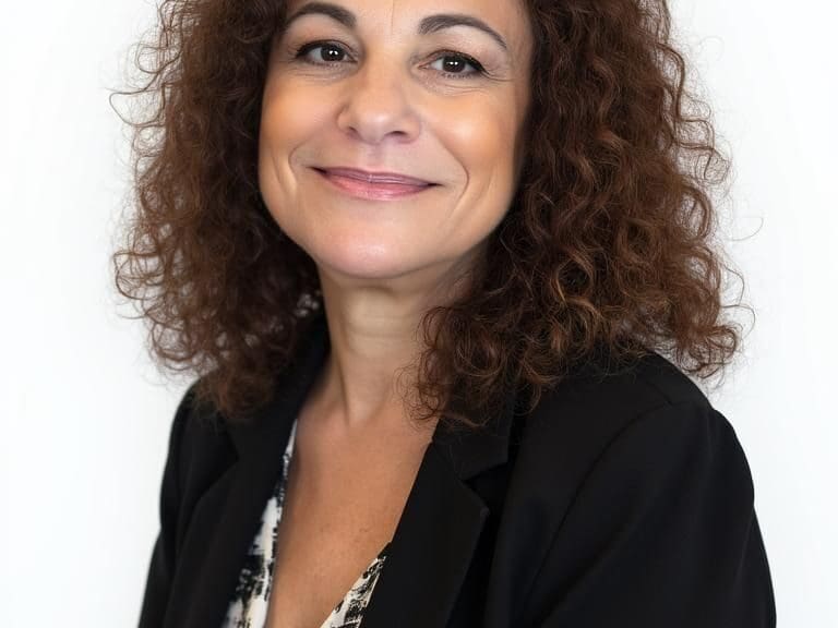 Portrait of a smiling woman with curly brown hair wearing a black blazer and a patterned blouse against a white background.
